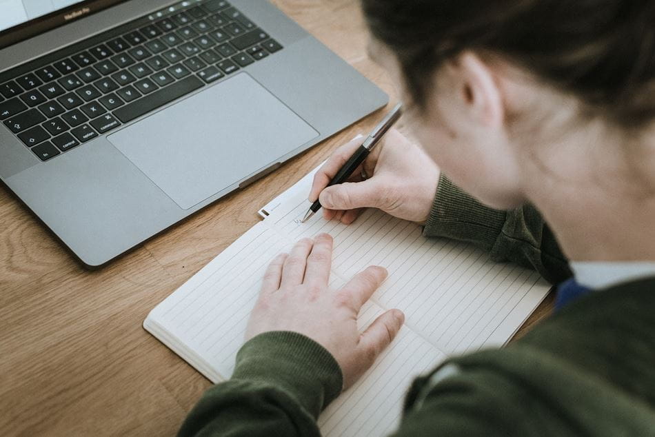 person writing on a notebook with a laptop open in front