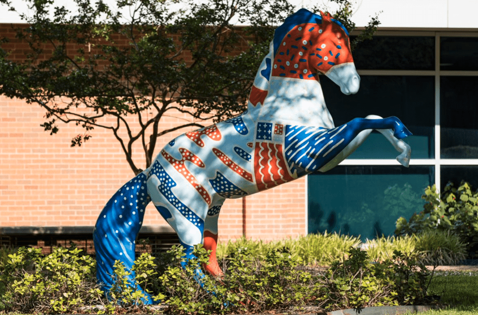 horse statue with blue tail surrounded by greenery in front of brick and glass building