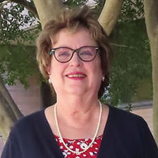 picture of Joyce Myers, dressed in a cardigan over a floral shirt with a pearl necklace, earrings, and glasses
