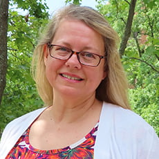 picture of LaDonna Aiken, dressed in a cardigan over a floral shirt with dark-rimmed glasses in front of trees on campus