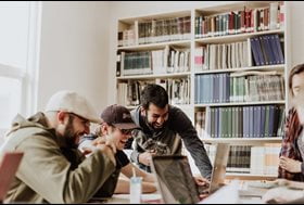 four students gathered around a table with their laptops, a bookshelf in the background