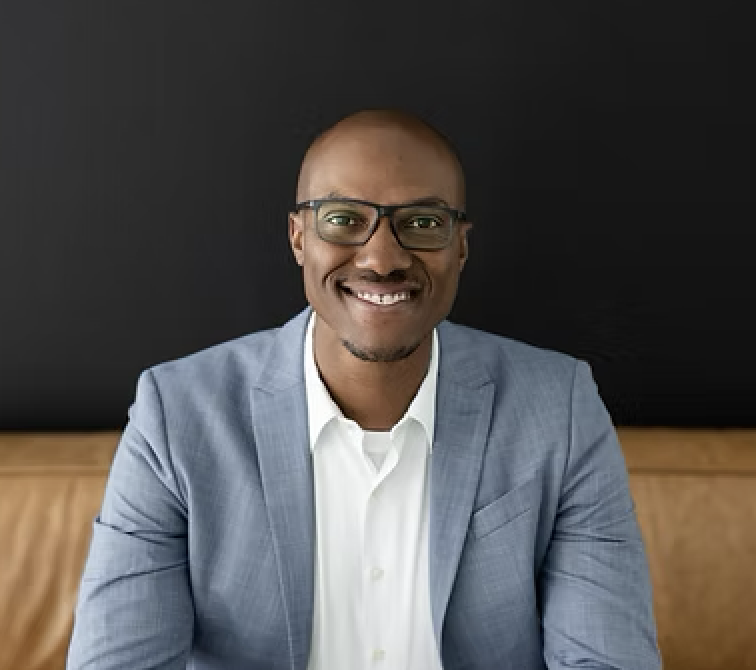 Nathan Hervey sitting on a couch, hands clasped together, smiling.