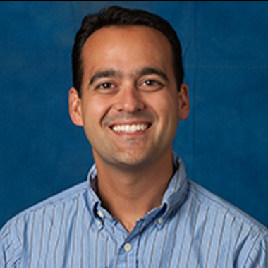 Steven Arxer in a dress shirt with short hair, smiling in front of a plain background