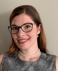 Eli Shupe smiling while wearing a dress blouse, glasses, and a pearl necklace.