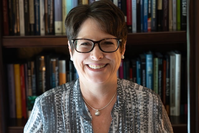 Miriam Byrd smiling at the camera, wearing glasses, earrings, necklace, and a patterned blouse.