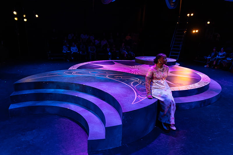 Woman in beautiful dress sitting on edge of pink and purple lighted stage