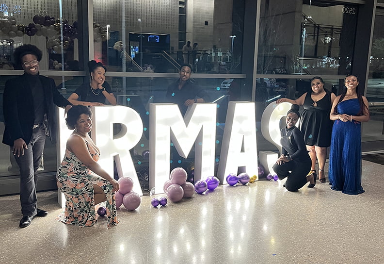 Students in formal attire stand in front of lighted Irmas sign.