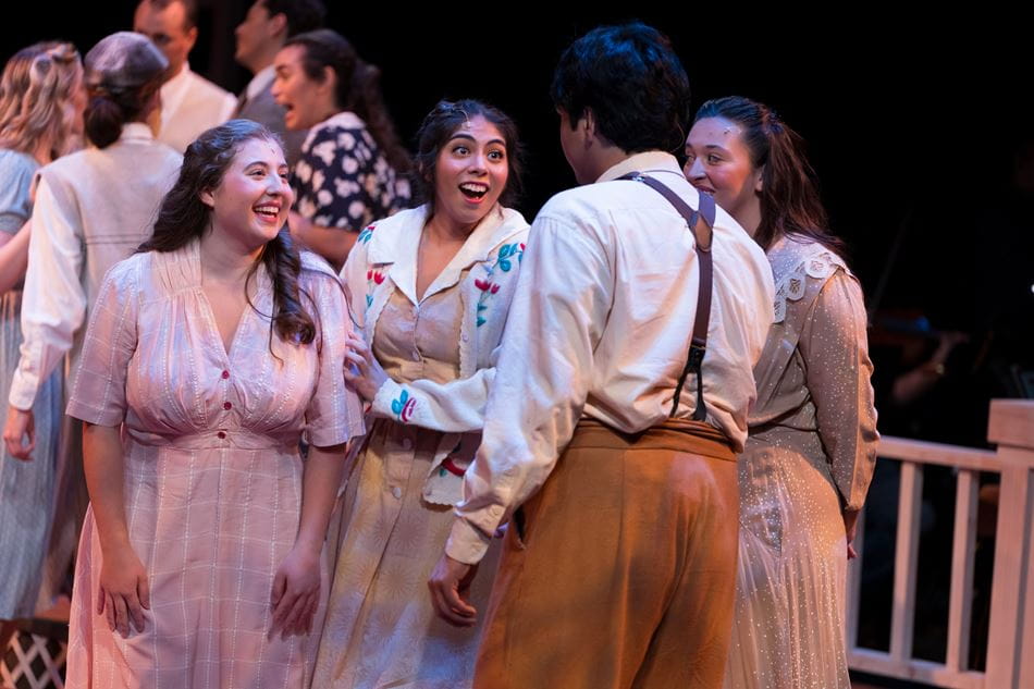 three women wearing period clothing on stage smiling at a man with his back to the camera