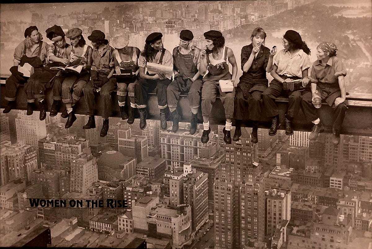 Sepia color of several women dressed as 1940-1950s construction workers sitting on steel beam with a cityscape below and behind them.  The title Women On the Rise is in the bottom left corner." _languageinserted="true