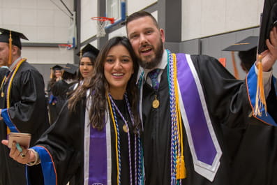 Two students wearing honors cords celebrate before a commencement ceremony.