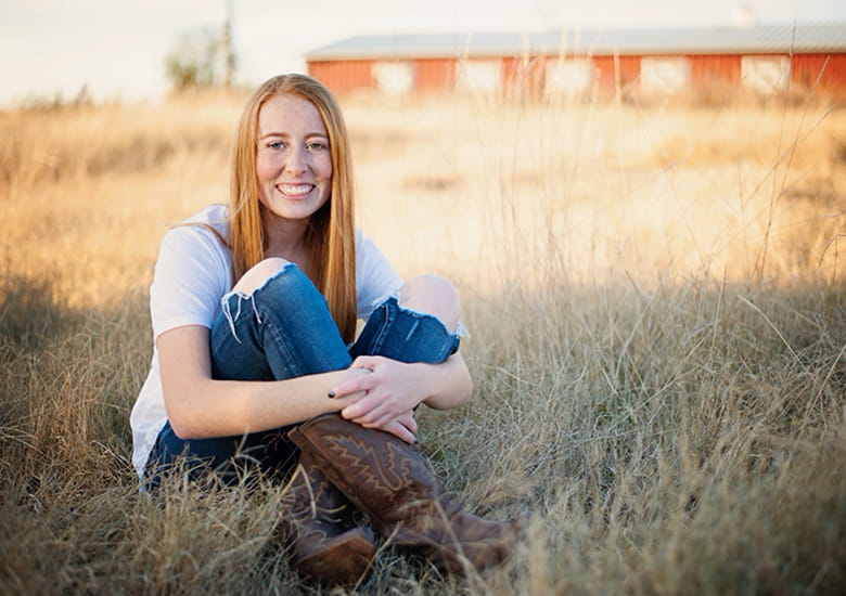 Aliza Capen wearing jeans and cowboy boots sitting in a large field with a barn in the background