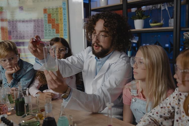 A teacher uses beakers to explain a science project. Students are wearing goggles and a periodic table of elements is visible in the classroom.