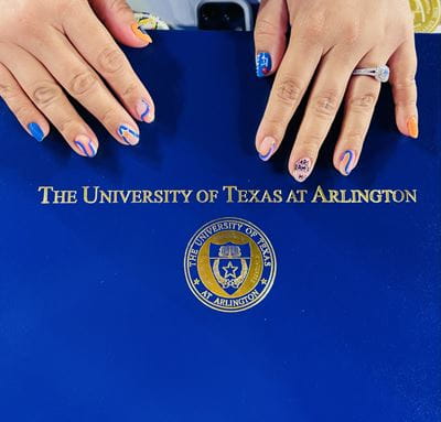 Photo of a student showing off blue and orange nails (the UTA colors) while holding the outside of a diploma cover reading "The University of Texas at Arlington" 