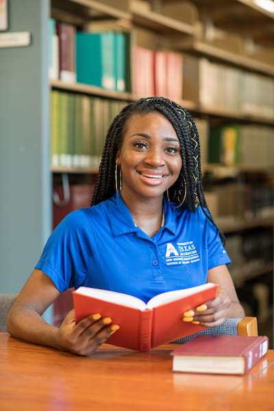 Photo of a UTA student in the library smile while holding a book that she's been reading