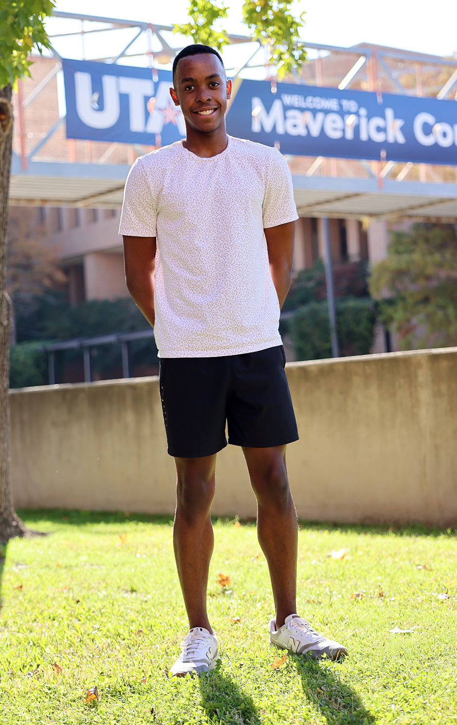 UTA bioengineering student Andrew Hackshaw standing in grass with the Cooper St. pedestrian overpass in the background with a "Welcome to Maverick Country" banner on it.