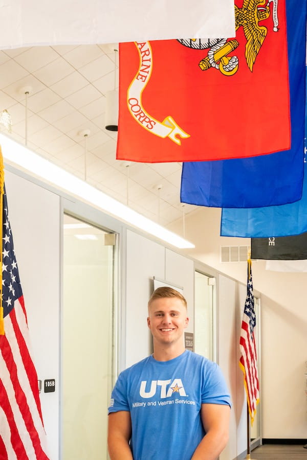 photograph of a man in a blue u t a military and veterans services shirt with a united states marine flag over head