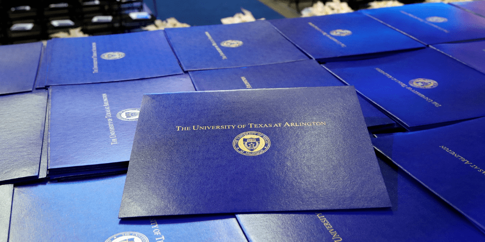 A stack of blue folders with the words The University of Texas at Arlington line a table at a graduate ceremony." _languageinserted="true