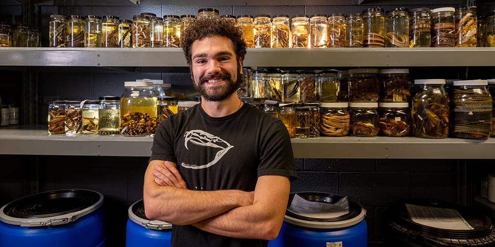 Greg Pandelis stands in front of shelves of jarred reptiles at the Amphibian and Reptile Diversity Research Center." _languageinserted="true