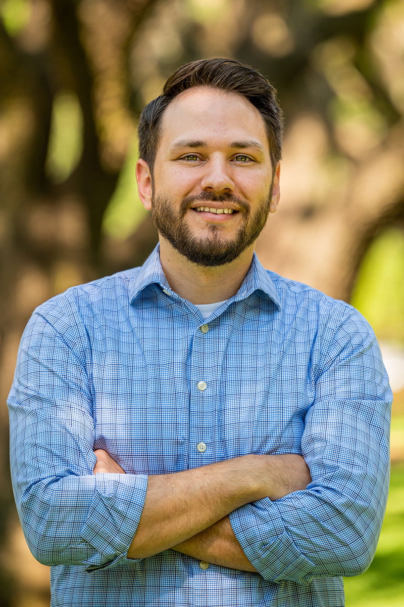 A man in a blue plaid shirt stands in front of trees and a green landscape." _languageinserted="true