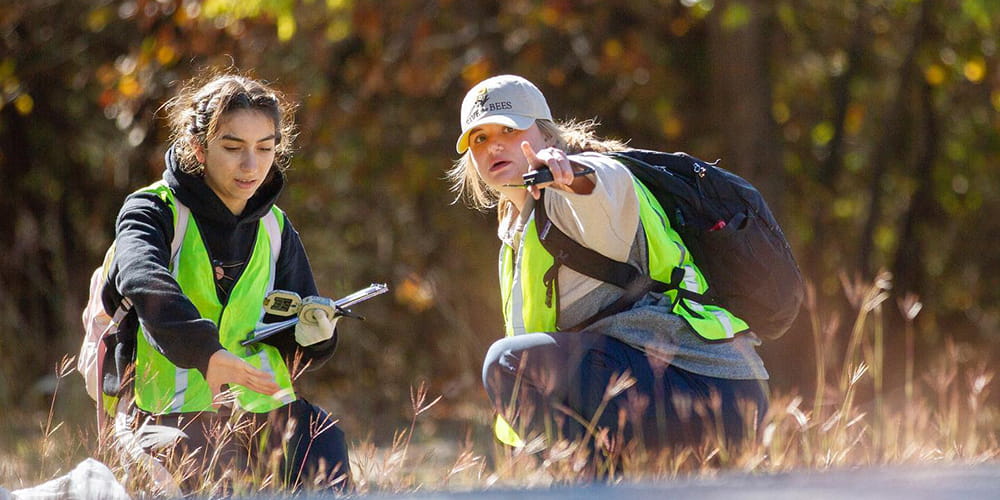 Two students kneel in a field where they are conducting research." _languageinserted="true