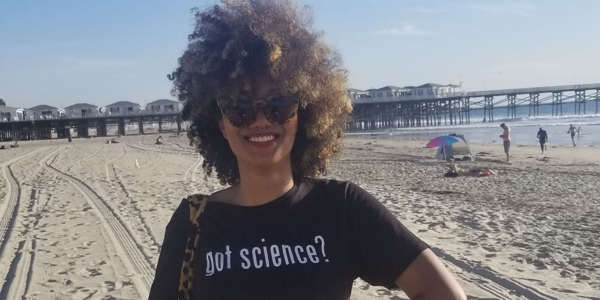 A young woman with brown curly hair stands on a beach. Her shirt reads, "got science?"" _languageinserted="true