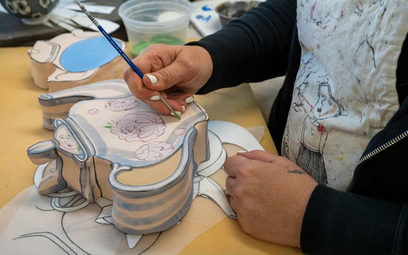 Яна Паюсова держит кисть и рисует фигурку чайника