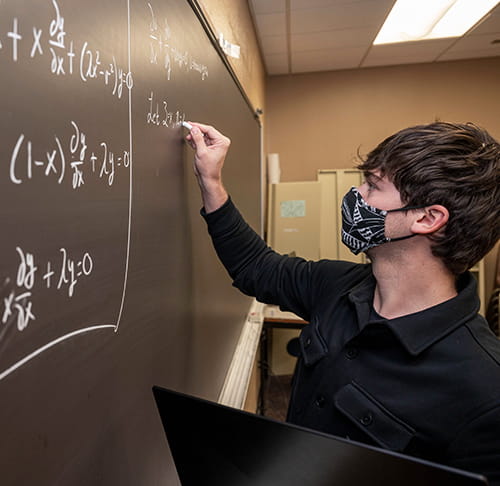 A male student wearing a mask writes math equations on a chalkboard.