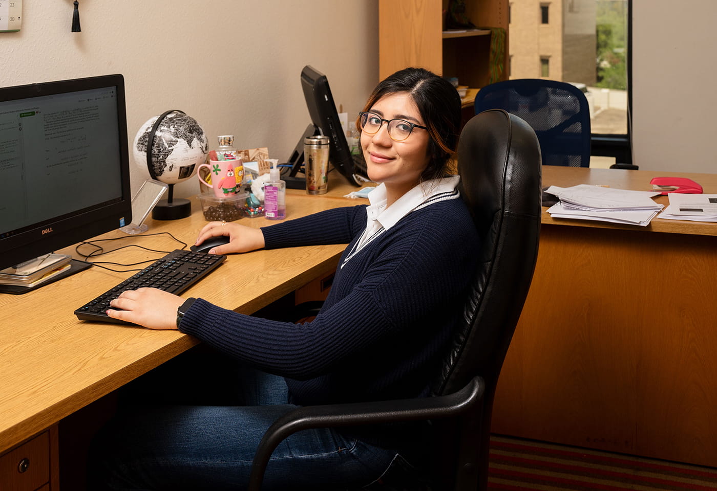 A young woman sits in front of a computer in a large office." _languageinserted="true