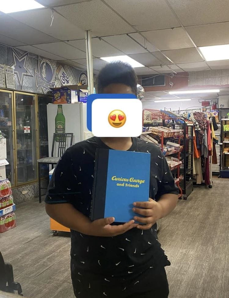 A kid holding a Curious George book with a smiley face emoji covering his face