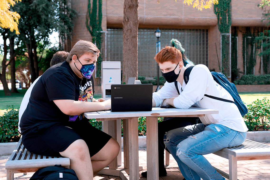 Students on a bench with face masks looking at a laptop