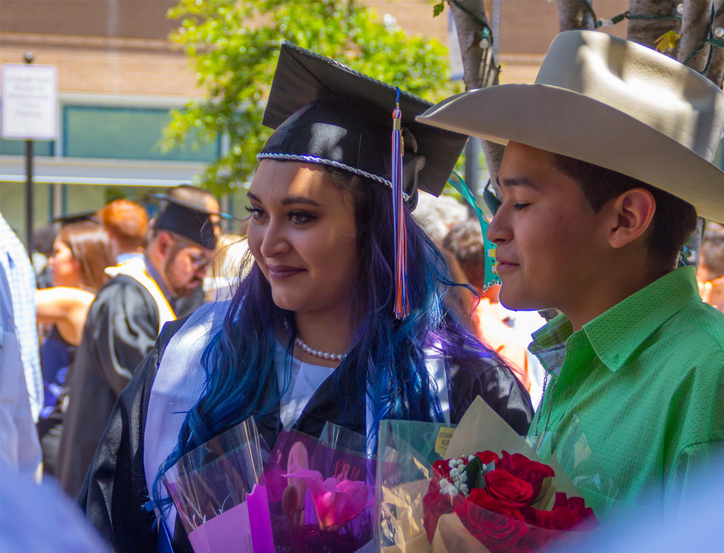 Woman with purple and blue hair in a graduation cap and gown posing with a boy in a cowboy hat.