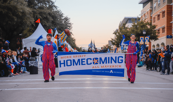 Two UTA students walk in the Homecoming parade while holding a Homecoming banner