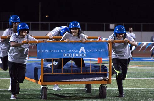 4 students run down Maverick Stadium rolling a bed on wheels with one student lying on it