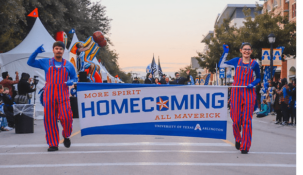 Two ambassadors in blue and orange stripes walking down the street with a sign between them labelled 'HOMECOMING'.
