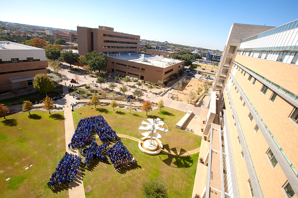 A group of UTA Students in blue shirts standing together to make the UTA logo