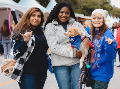 Three female students in warm clothing pose together with their golden retriever puppy sporting UTA attire