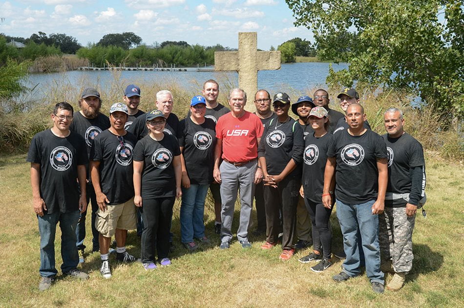 Group of men standing in front of a large cross sculpture outside near lake